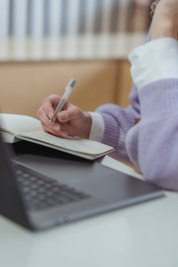 Crop faceless female freelancer working on netbook while writing in planner while sitting at table during remote occupation